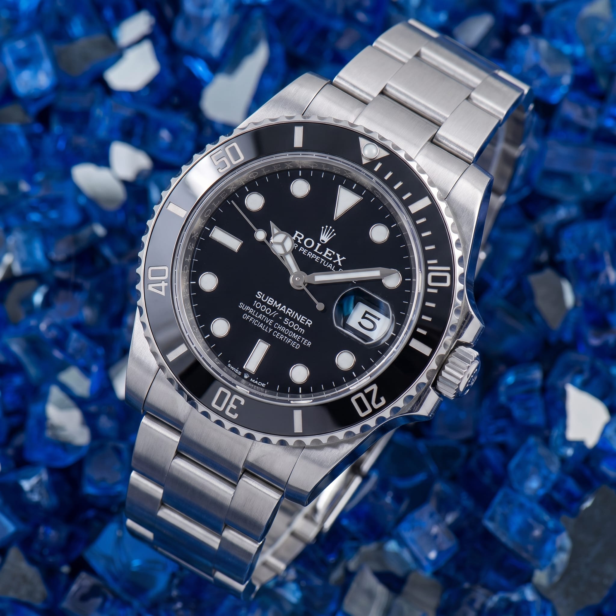 A genuine Rolex Submariner Date with a black dial and black Cerachrom bezel on an Oyster bracelet, shown dial-forward on a blue crushed-glass background